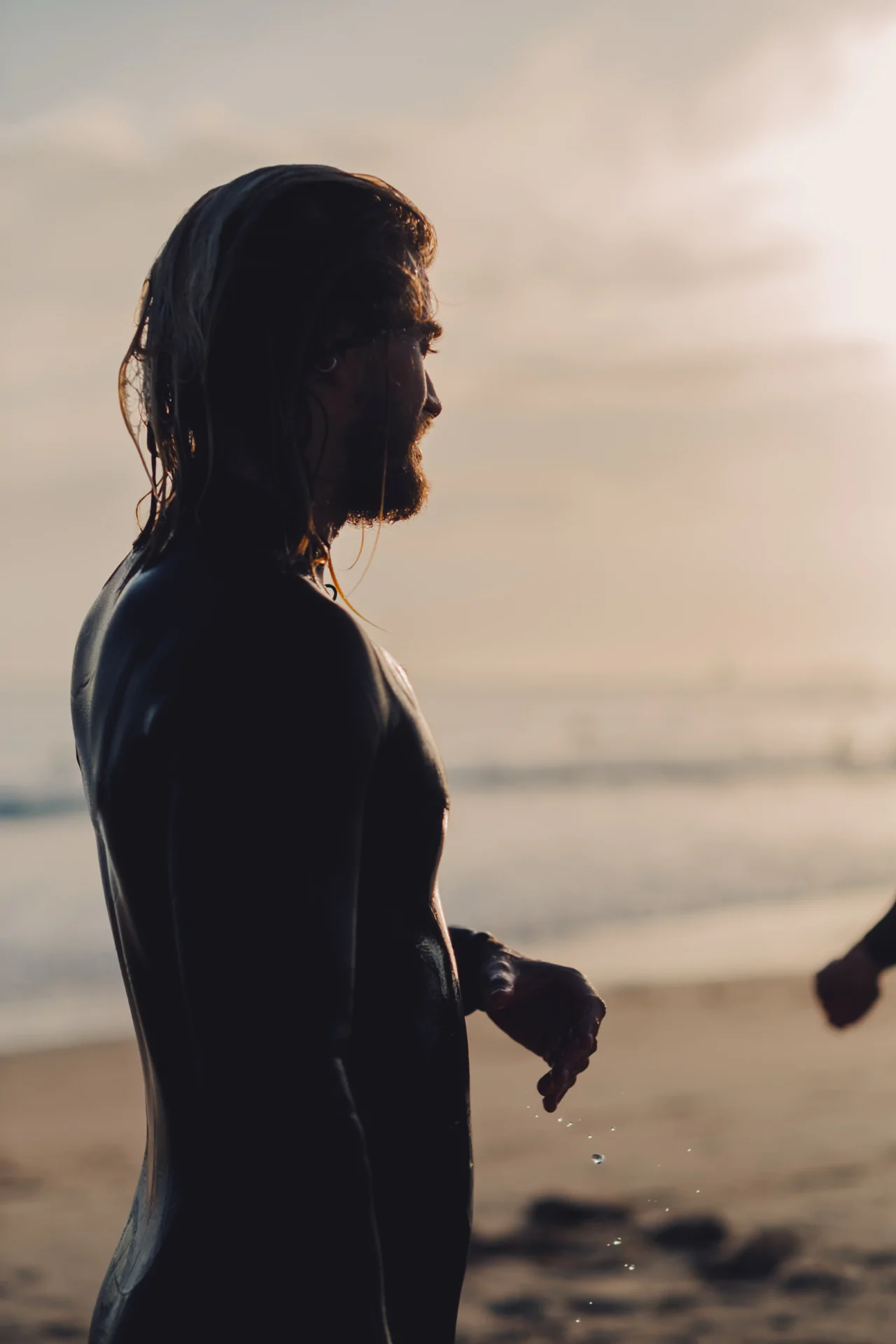 A person in a wetsuit stands on a beach at sunset, with wet hair and water droplets visible on their arm.