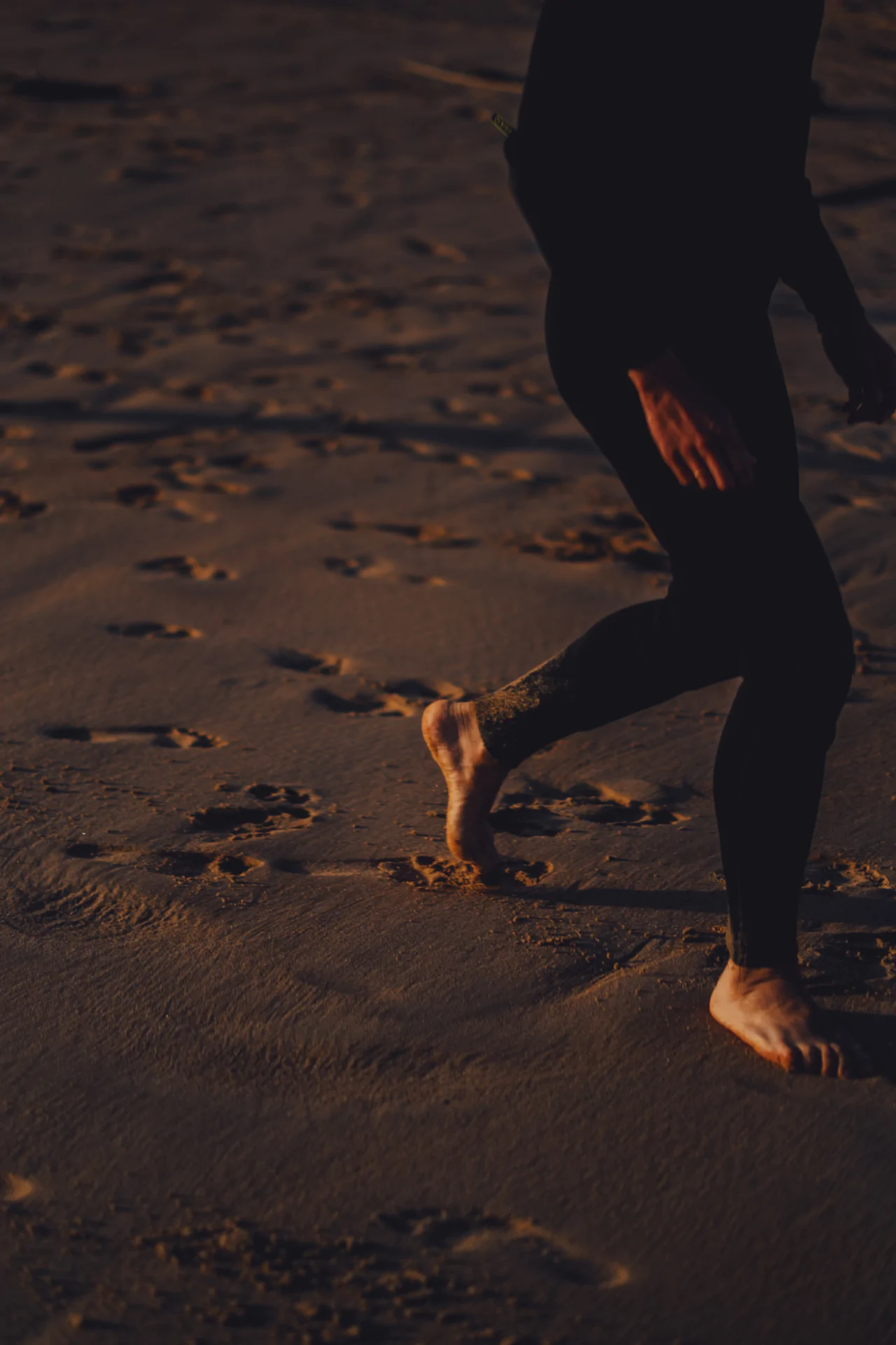 Person in a wetsuit walking barefoot on wet sand, with footprints visible in the evening light.