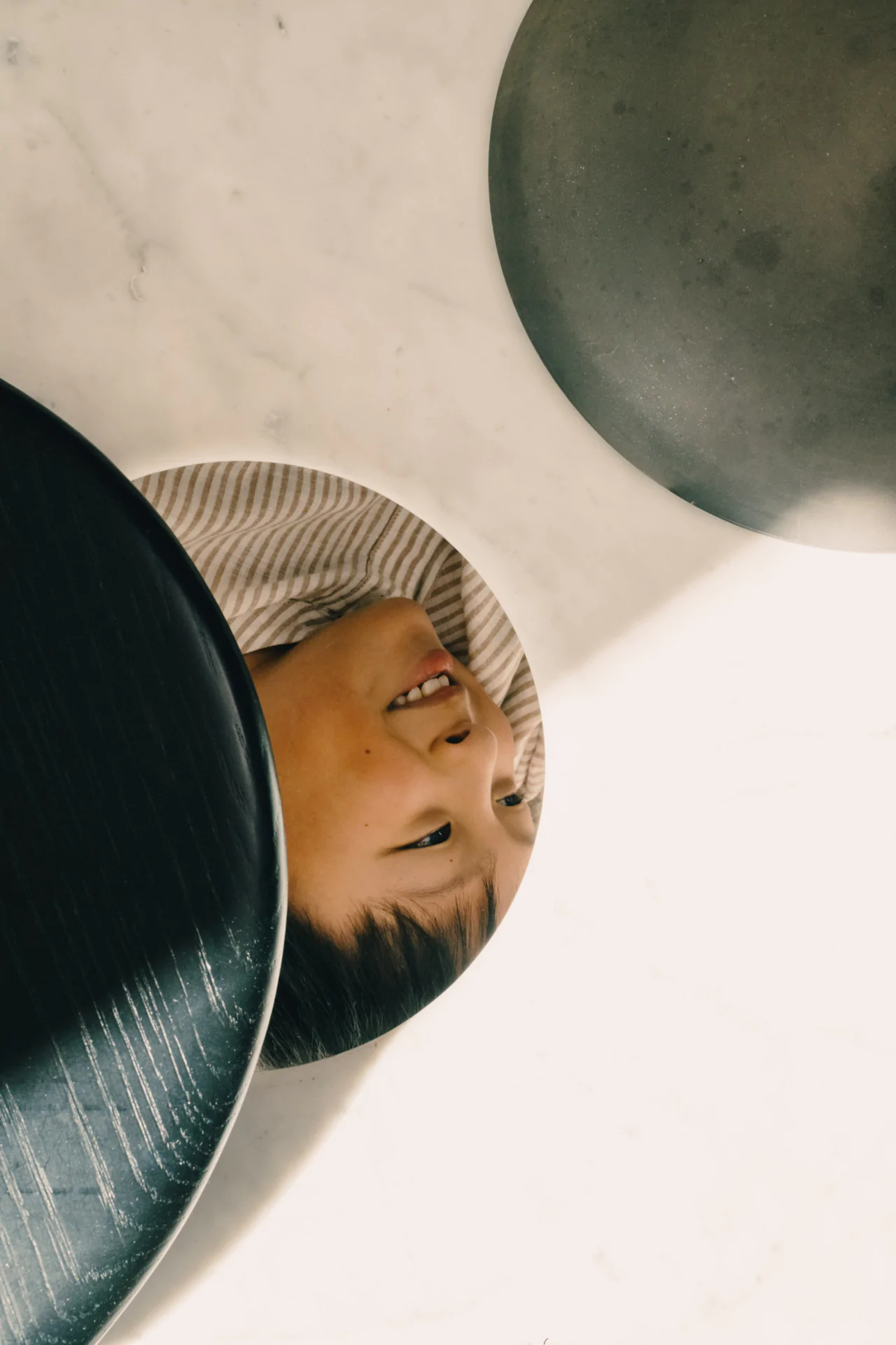A child is partially visible through a circular opening in a white surface, surrounded by dark round objects, smiling and looking up.