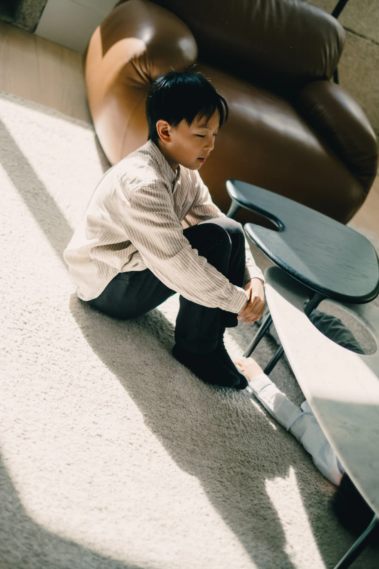A boy sits on a light-colored carpet next to a black table, with another child's legs visible beneath the table in a sunlit room.