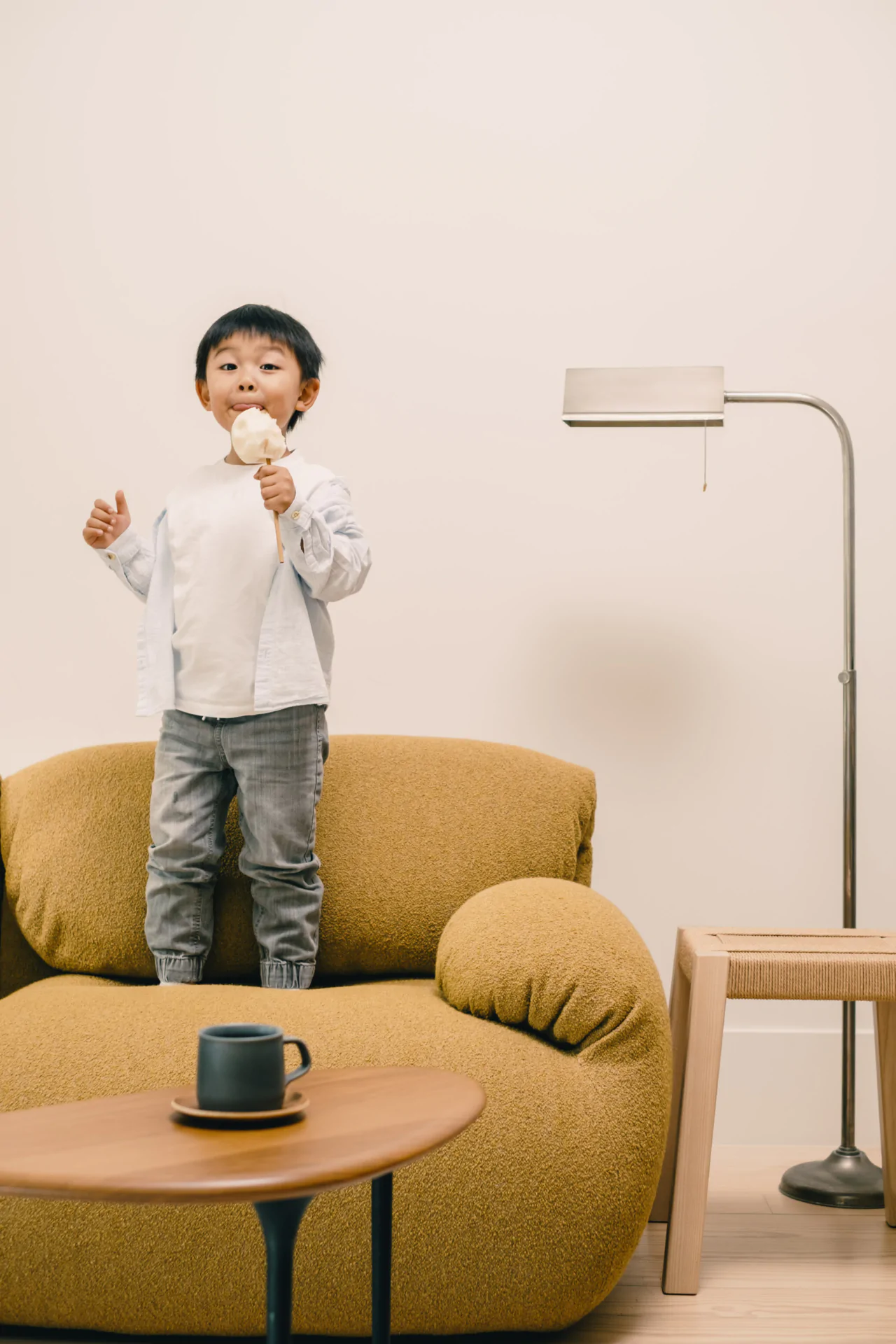 A young boy stands on a mustard-yellow sofa holding an ice cream cone, with a floor lamp, side table, and a coffee cup in the background.