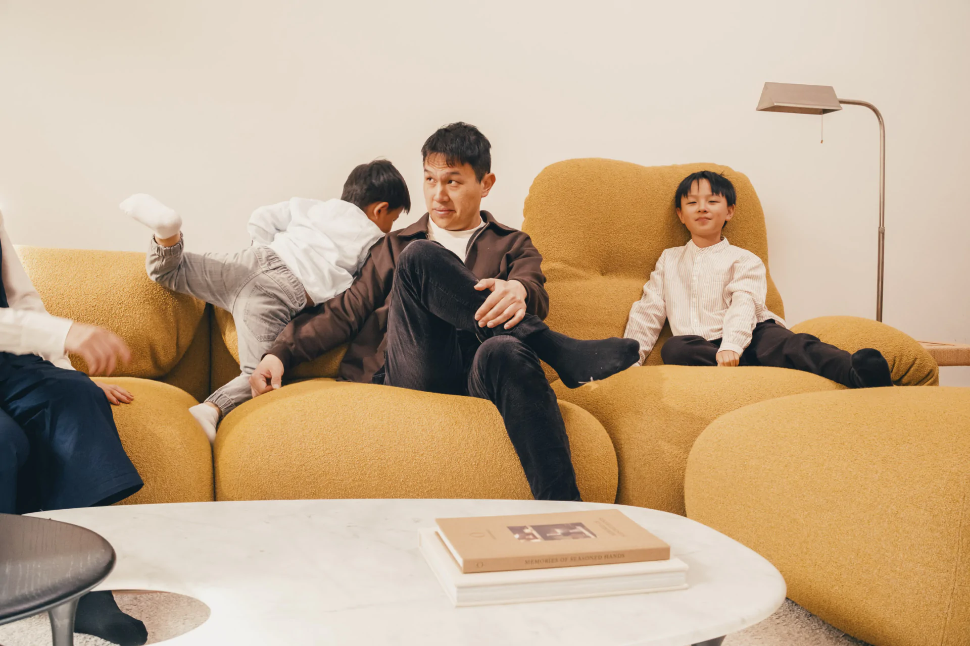 A man and two children sit on yellow couches; one child is climbing over the man while the other sits calmly. A round white table with books is in the foreground.