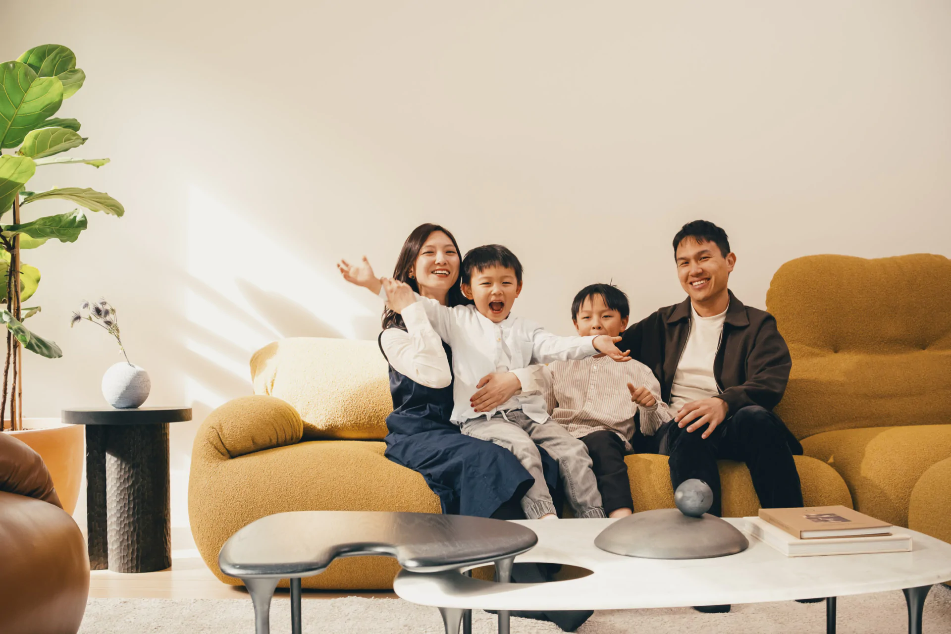 A family of four sits together on a yellow sofa in a living room, smiling and appearing cheerful. A plant and coffee table with books are visible in the foreground.