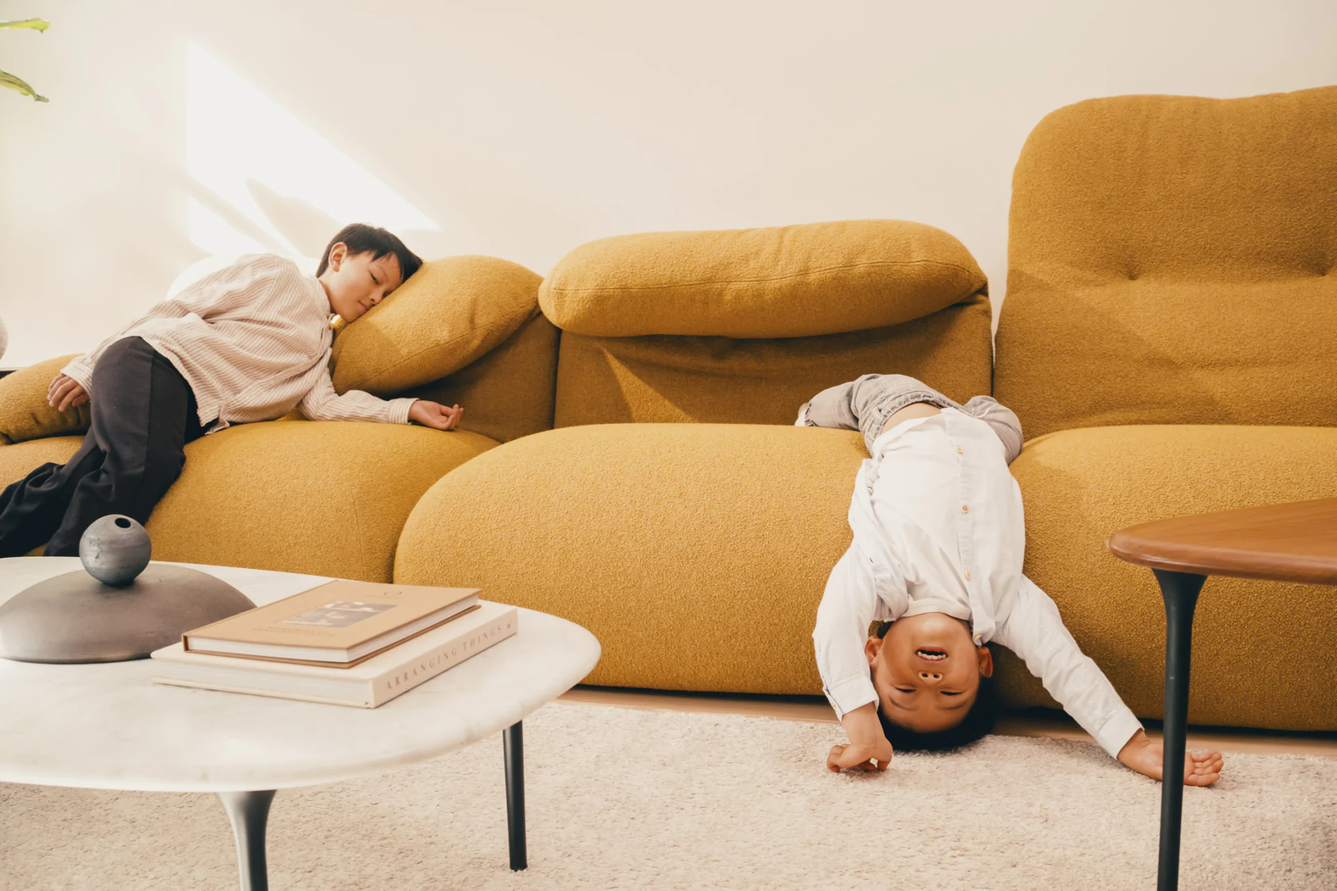 Two young children relax on a mustard-colored sofa; one lies down while the other hangs upside down, feet up and head near the floor. A coffee table with books is in the foreground.