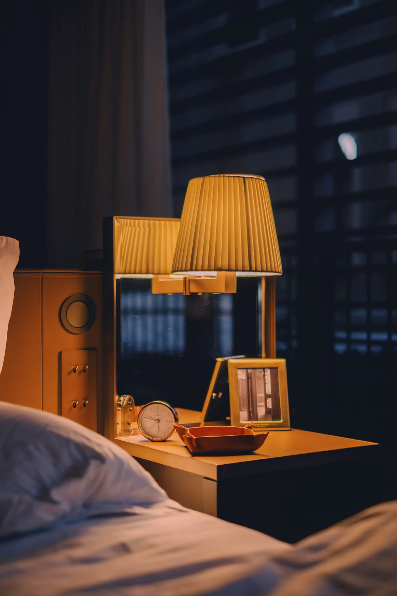 A bedside table with a lamp, a framed photo, an analog clock, a tray, and a mirror next to a bed at night, captured in the style of luxury hotel photography São Paulo.