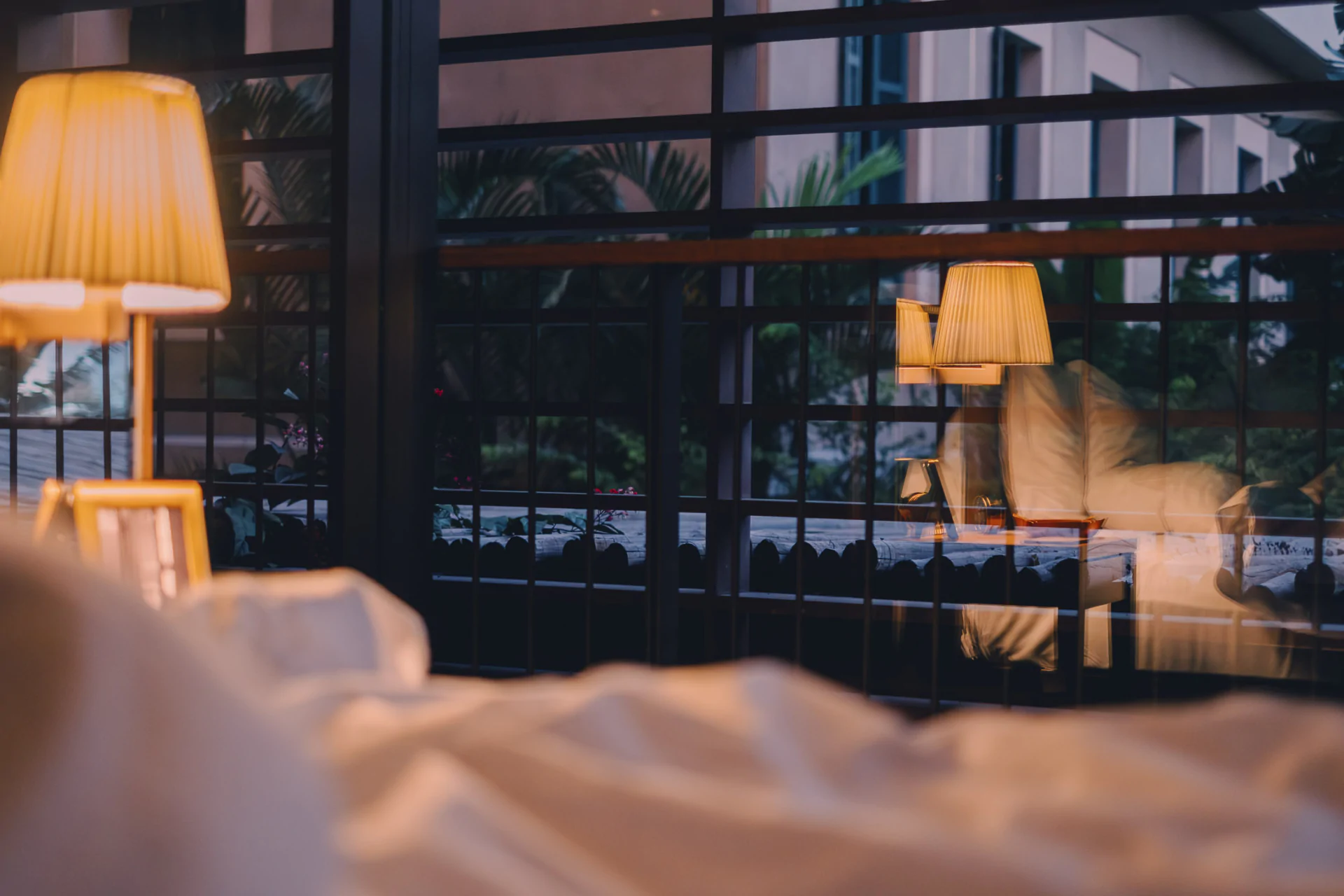 A bedside lamp glows beside a bed, its reflection visible in a large window showing greenery and part of a building outside.