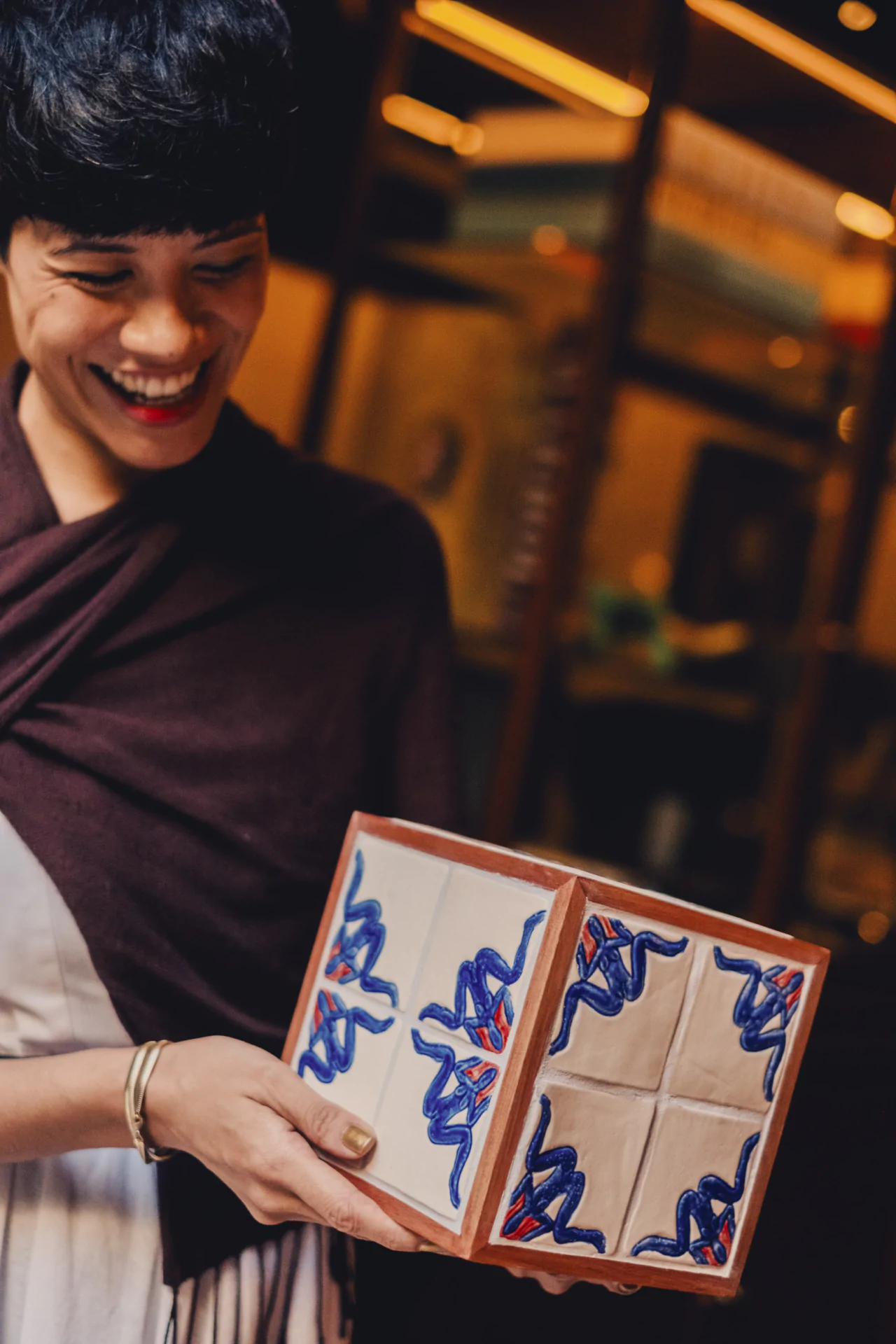 A person smiles while holding a ceramic cube decorated with abstract blue and red patterns, standing indoors.