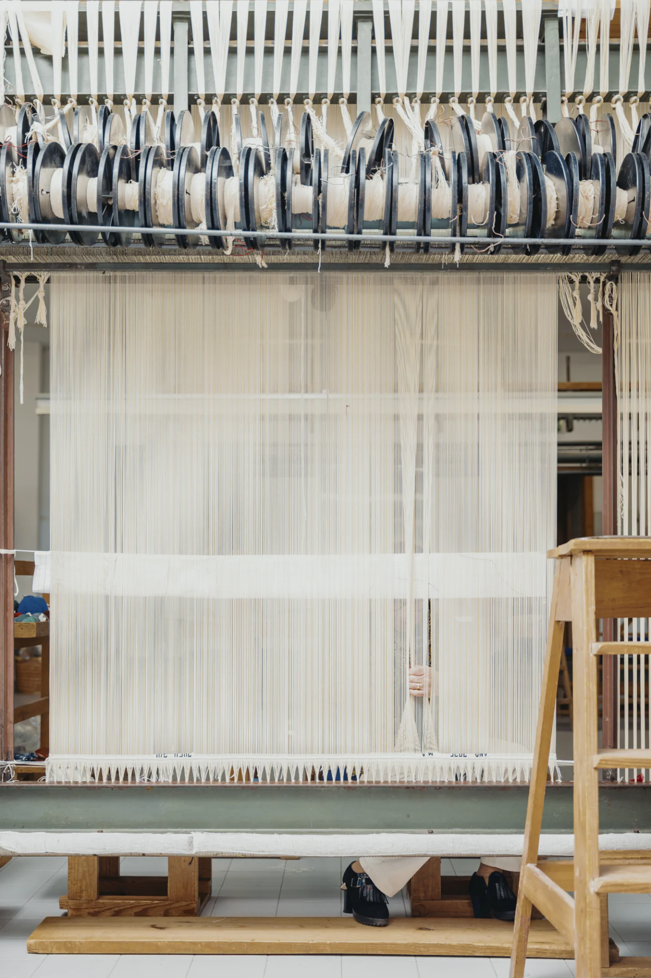 A person operates a large loom, with threads strung vertically across the frame and their hands visible adjusting the threads from behind the machine.