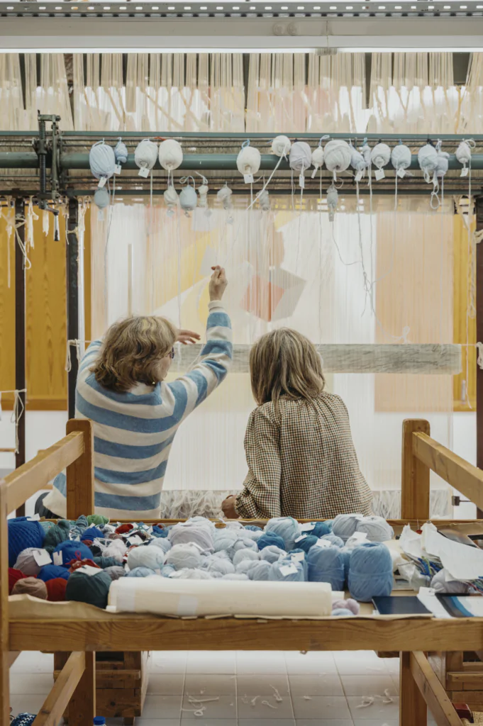 Two people sit at a loom, weaving with yarn and threads. Various balls of yarn are arranged on the table in front of them.