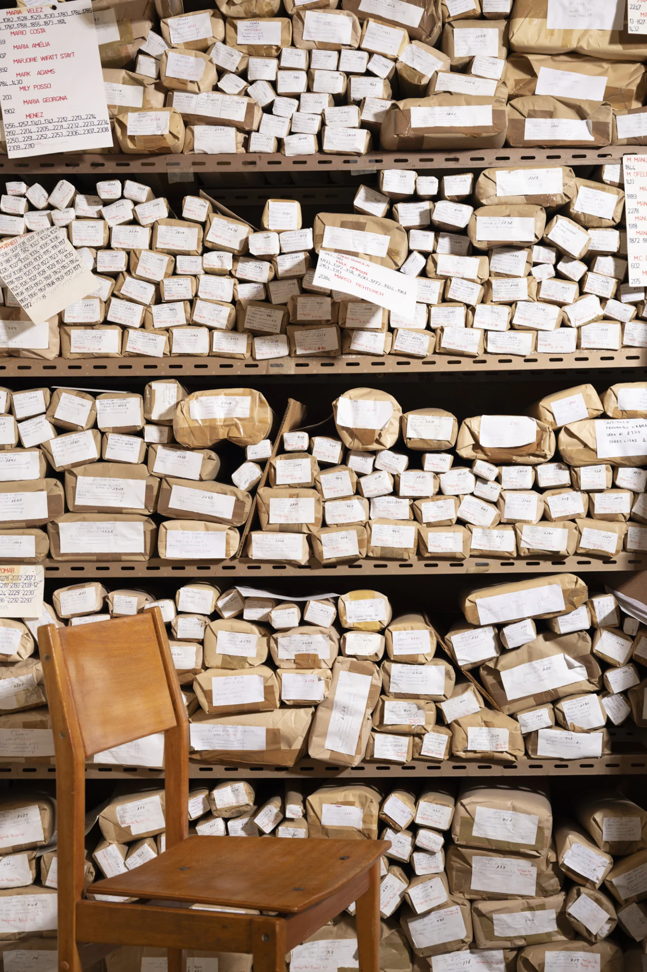 Wooden chair in front of shelves stacked with numerous brown paper-wrapped parcels, each labeled with handwritten notes. Various tags and paper signs are attached to the shelves.