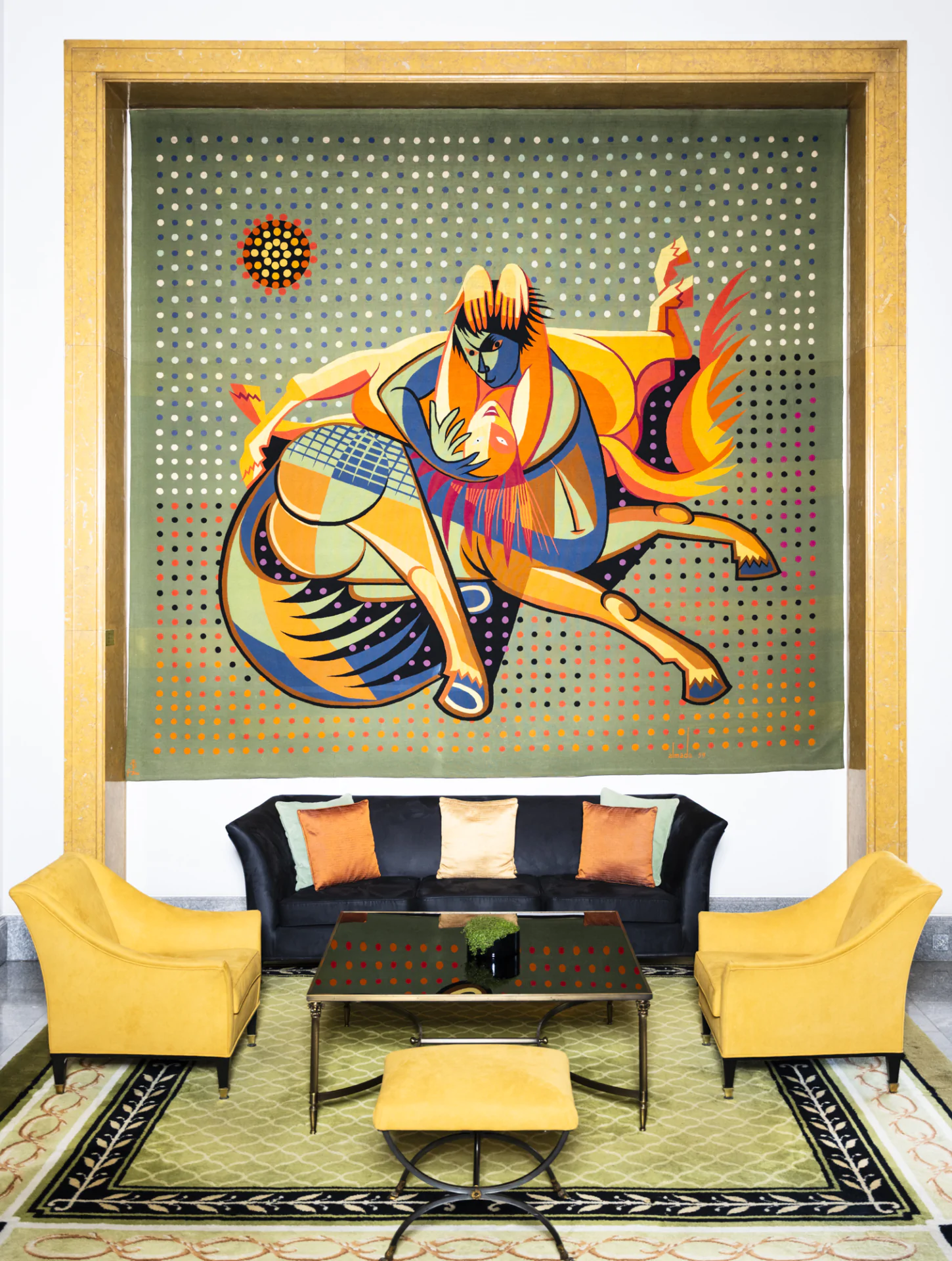 A modern lounge with yellow armchairs, a black sofa with colored cushions, a glass coffee table, and a large abstract tapestry of a horse and figure on the wall.